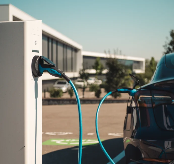 Vue rapprochée d'une station de recharge connectée Nexteneo avec un cordon électrique à une voiture électrique. En arrière-plan, un parking équipé d'un parc de stations de recharge