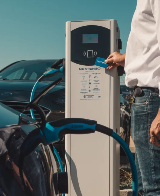 Homme utilisant une carte pour activer une station de recharge connectée Nexteneo en plein air, avec une voiture électrique en arrière-plan.